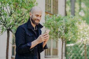 Positive bald young male with stubble, holds modern cell phone, enjoys online communication in social networks, has glad expression, dressed in black jacket, poses outdoor against green trees photo