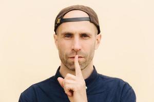 Close up portrait of attractive young male gestures indoor, shows silence sign, has serious expression, wears stylish cap, poses against beige background. People, secrecy and conspiracy concept photo