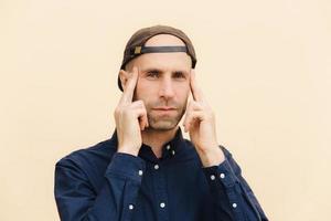 Indoor shot of attractive male keeps fingers on temples, tries to concentrate on some problem, recollects important information in mind, poses against blank studio wall. People and facial expressions photo