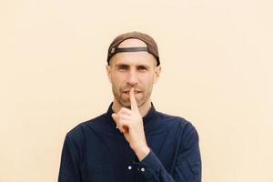 Horizontal shot of handsome young male model keeps fore finger on lips, asks to be quiet, wears cap and shirt, poses against beige background. Secret Caucasian man gestures indoor. Hush sign photo