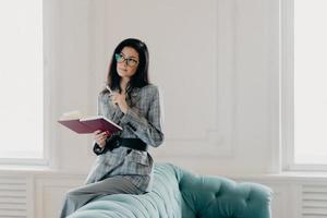 Serious businesswoman dressed in formal wear, concentrated into distance, ponders on creative ideas, writes down notes in notepad, wears spectacles, poses on back of couch in spacious light room photo