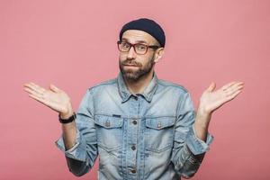 Indoor shot of hesitant bearded man shrugs shoulders, looks uncertain, doubts when makes decision, isolated over pink background. People, doubt and hesitation concept. Body language concept. photo