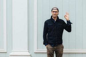 Indoor shot of handsome cheerful unshaven male shows okay sign, wears stylish shirt, cap and trousers, keeps hand in pocket, poses against wall, satisfied with something, Body language and people photo