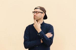 Indoor view of attractive middle aged fashionable male dressed in shirt and cap, looks pensively aside, thinks about something, poses against beige background. People, lifestyle, thoughts concept photo