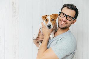 Horizontal portrait of handsome cheerful man, wears eyeglasses, holds jack russell terrirer, has glad expression, poses against white wooden wall with blank copy space. Animals and friendship photo