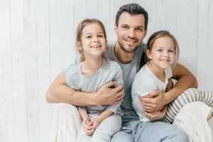 retrato de padre joven feliz abraza a sus dos hijas, las ama mucho, posan juntos en casa. el padre soltero con hermosas niñas tiene expresiones alegres. concepto de paternidad foto