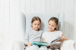 dos hermanitas adorables se sientan en el sofá, leen un libro interesante, se sientan en un cómodo sofá, disfrutan de la unión. las mejores amigas tienen un libro, ven fotos y leen a los oyentes, tienen tiempo libre