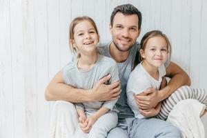 padre e hijos. concepto de paternidad. apuesto padre cariñoso sin afeitar abraza a sus dos lindas hijas, van a caminar juntas, tener una buena relación, posar en la cámara con sonrisas positivas foto