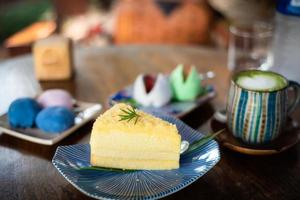 pastel de gasa de queso crema de primer plano en un plato de baldosas decorado con hojas de romero y té verde matcha encima arte latte decorado con espuma de leche en forma de corazón en una taza colorida, pastel de estilo japonés foto