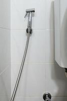 The metal bidet spray is hanging on the white tile wall near the flush toilet. photo