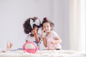 A cute little Asian girl and African friend is happily applying makeup brushes with powder in her bedroom photo