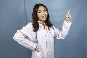retrato de una joven doctora asiática, un profesional médico sonríe y señala hacia arriba en un espacio de copia aislado sobre fondo azul foto