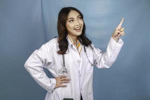 Portrait of a young Asian woman doctor, a medical professional is smiling and pointing upward at a copy space isolated over blue background photo