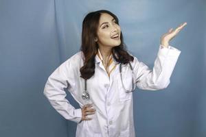 Portrait of a young Asian woman doctor, a medical professional is smiling and pointing upward at a copy space isolated over blue background photo