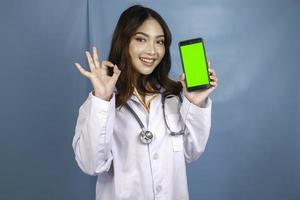 una joven doctora asiática está señalando y mostrando una pantalla verde o copiando espacio en su teléfono inteligente. mostrando los pulgares hacia arriba o el signo de ok. foto