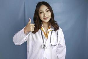 Portrait of a young Asian woman doctor, a medical professional is smiling and showing thumbs up or OK sign isolated over blue background photo