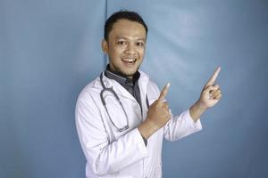 Happy young Asian man doctor, a medical professional is smiling and pointing upward at a copy space isolated over blue background photo