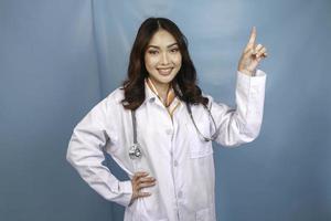 Portrait of a young Asian woman doctor, a medical professional is smiling and pointing upward at a copy space isolated over blue background photo