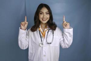 Portrait of a young Asian woman doctor, a medical professional is smiling and pointing upward at a copy space isolated over blue background photo