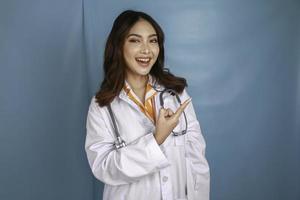 Portrait of a young Asian woman doctor, medical professional is smiling and pointing at a copy space isolated over blue background photo