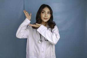 Portrait of a young Asian woman doctor, a medical professional is smiling and pointing upward at a copy space isolated over blue background photo