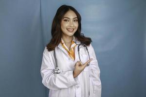 retrato de una joven doctora asiática, profesional médico sonríe y señala un espacio de copia aislado sobre fondo azul foto