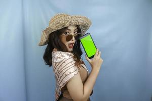 Young Asian happy woman showing green screen on her smartphone isolated by a blue background photo