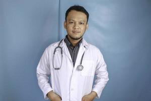 Confident relaxed male Asian doctor standing with hands in the pockets of his lab coat smiling quietly at the camera over blue background with copy space photo