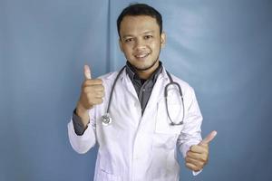 retrato de un joven médico asiático, un profesional médico sonríe y muestra el pulgar hacia arriba o el signo ok aislado sobre fondo azul foto