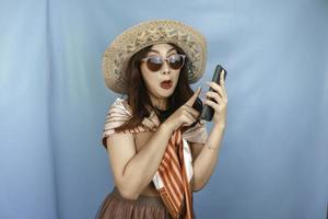 Young Asian woman pointing and shocking to her smartphone isolated by a blue background photo