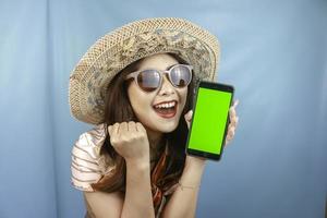 Young Asian happy woman showing green screen on her smartphone isolated by a blue background photo