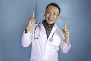 Happy young Asian man doctor, a medical professional is smiling and pointing upward at a copy space isolated over blue background photo