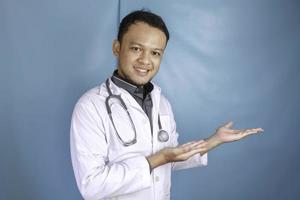 Happy young Asian man doctor, medical professional is smiling and pointing at a copy space isolated over blue background photo