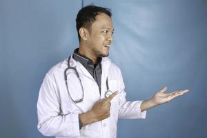 Happy young Asian man doctor, medical professional is smiling and pointing at a copy space isolated over blue background photo