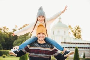 el retrato horizontal de un padre afectuoso le da un paseo a cuestas a su hija afuera, juegan juntos, se divierten. feliz niñita encantadora pasa los fines de semana con su padre, saluda emocionada con la mano foto