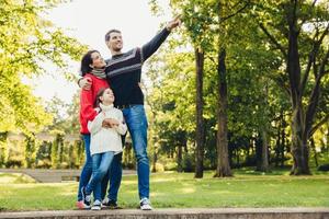 Image of happy joyful young family father, mother and little daughter play together in autumn park, countryside, enjoy nature outside. Attractive man shows something with hand daughter and wife