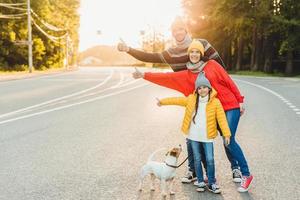 Carefree relaxed friendly family walk on road in countryside with dog, raise thumbs with enjoyment, wear warm clothes and sport shoes as have long way. Young family gestures as has free time photo