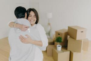 Happy brunette smiling woman embraces with love her husband, holds keys, buy first house together, purchase new property, pose in unfurnished room with pile of cardboard boxes. Ownership concept photo