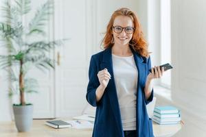 una próspera y sonriente banquera pelirroja paga el servicio en línea, espera una llamada importante, sostiene un teléfono celular moderno, mira positivamente a la cámara, se viste con ropa elegante, posa en la oficina foto