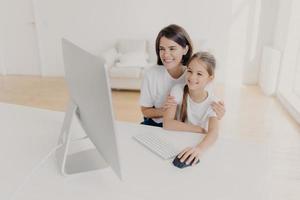 Happy brunette mother helps child do homework on computer, sit together at workplace, have cheerful expressions, focused into monitor. Little girl plays video games via modern gadget with mom photo