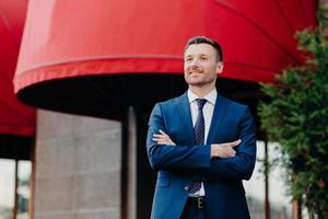 Self assured male CEO in formal suit stands crossed hands, looks with satisfied expression into distance, waits for colleagues near cafe, going to have corporative meeting, thinks about work photo