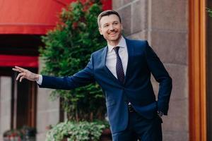 Horizontal shot of happy male manager makes hitchhike gesture, dressed in formal clothing, stands near cafe, asks taxi to give him lift, has happy expression. People, business and positiveness photo