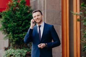 Successful businessman has work break, uses mobile phone for having talk, being always on line, dressed in formal suit, has pleasant smile on face, stands outdoor near restaurant. Technology concept photo