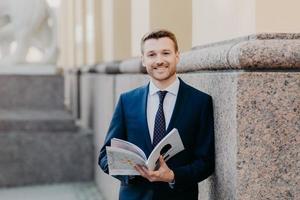 un próspero y exitoso hombre de negocios vestido formalmente lleva un diario, tiene una expresión feliz, sonríe alegremente, disfruta de la cena después de un duro día de trabajo, está de buen humor después de una reunión de negocios foto