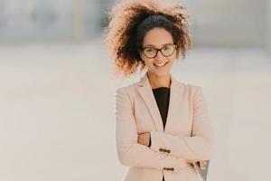 Confident cheerful female entrepreneur with curly Afro hair, keeps arms folded, uses modern tablet computer, smiles positively, waits for business partner outdoor, blurred urban setting in background photo