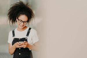 Beautiful African American girl in fashionable clothing, uses mobile phone for sending text messages, connected to wifi, checks email box, stands near white wall with empty space. Online communication photo