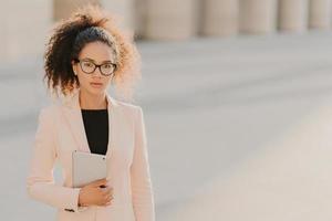 Photo of elegant African American businesswoman uses tablet computer outdoor, wears formal white jacket, transparent glasses with black frames, curly hair combed in pony tail, blank space on left