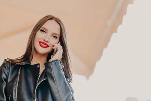 Shot of beautiful young millennial woman enjoys talk with boyfriend, wears leather jacket, has makeup, red lipstick, uses modern cell phone for communication. People and conversation concept