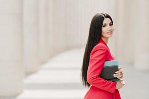 Sideways shot of elegant brunette lady with dark long straight hair, dressed in fashionable red suit, has manicure, carries diary, stands outdoor over white building. Female employee in formal apparel photo