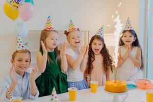 Cheeful friends of five pose near festive table with big cake, cup of drinks, wear party hats, look joyfully at sparkle, laugh happily, pose indoor, celebrate birthday photo